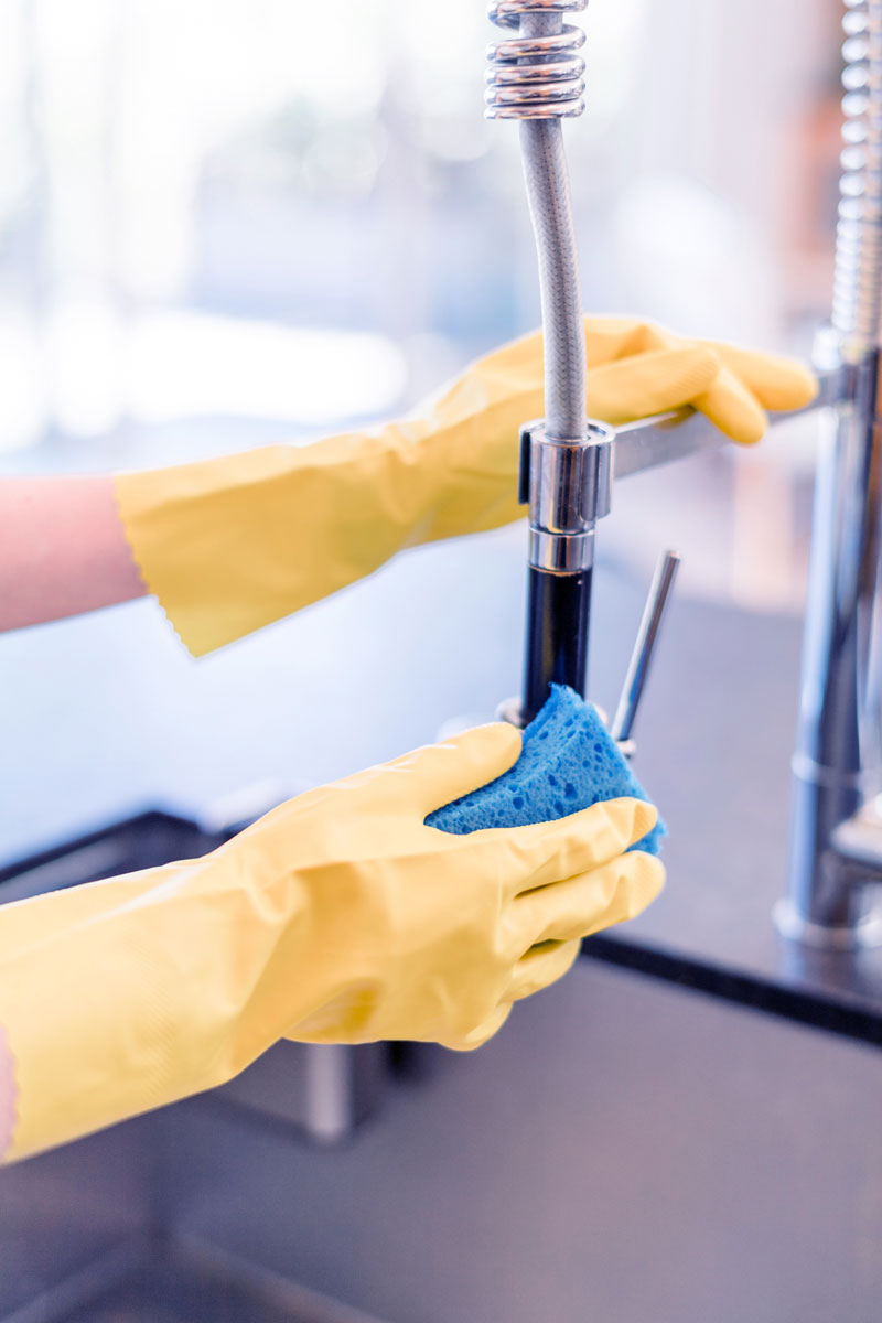 A person wearing yellow rubber gloves uses a blue sponge to clean a kitchen faucet.